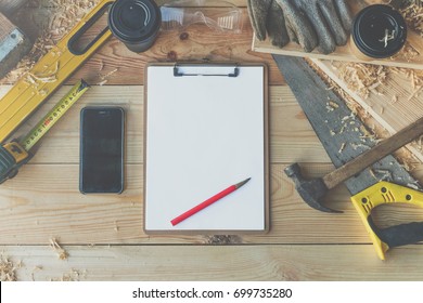 On Wooden Floor Is Clipboard With Empty White Sheet, Pencil.Nearby Is Smartphone,cup Of Coffee And Various Carpentry Tools: Roulette, Hammer, Saw, Level, Plane, Building Gloves, Wooden Sawdust.Mock Up