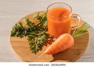 On a wooden kitchen board there is a glass with healthy carrot juice, fresh carrots with green leaves. 