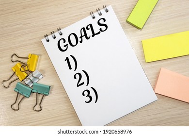 On the wooden table among the colored paper clips and stickers there is a notebook on which the goals are written with an enumeration.