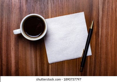 On a wooden table next to a white cup of coffee and a pen is a clean white paper napkin. Copy space
