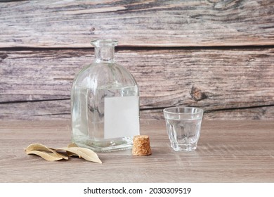 On a wooden table is an uncorked bottle of schnapps with a blank label and a full shot glass. The stopper of the bottle is next to it and there are also some dried laurel leaves. - Shutterstock ID 2030309519