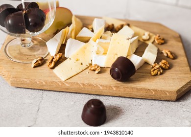 On a wooden tray lies cheese, chocolates, walnuts and chopped apples.