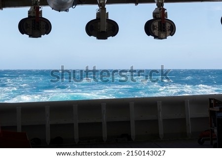 Onboard view from streamer deck towards the aft of seismic survey vessel. Empty cable blocks hanging from the top. Clear weather. Moderate sea state. Blue and turquoise water with white caps.