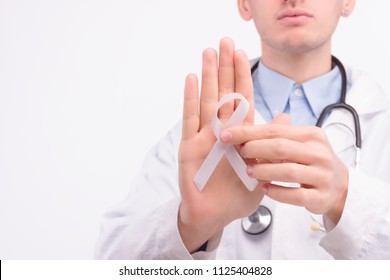 Oncological Disease Concept. Doctor Wearing White Coat And Stethoscope Putting White Ribbon As A Symbol Of Lung Cancer On His Right Hand.