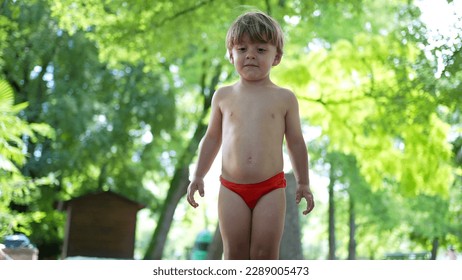 One adorable active small boy jumping into water puddle barefoot during summer day wearing child bathing suit briefs. Active kid splashing into park pool