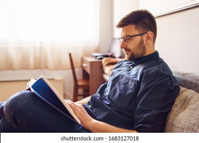 One adult caucasian man sitting on the sofa bed at home reading the book study learning alone during the quarantine lockdown in a day wearing shirt and eyeglasses and beard side view - Powered by Shutterstock