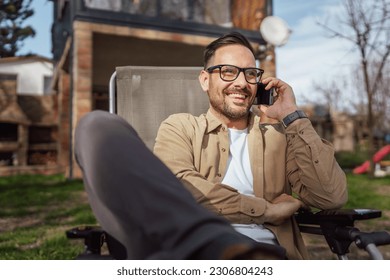 One adult man 40 years old caucasian male sit in front of his house on vacation talk on his smartphone mobile phone call happy smile during the conversation real person copy space - Shutterstock ID 2306804243