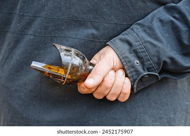 one aggressive man in a black jacket holds in his hand a piece of a broken sharp heavy dangerous brown glass bottle behind his back before an attack on the street 
