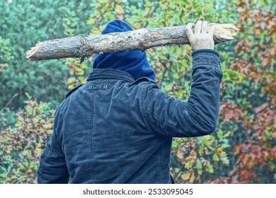 one aggressive man in a brown jacket and a blue hood on his head holds a heavy wooden wide gray stick in his hand and swings it before attacking during the day in the forest