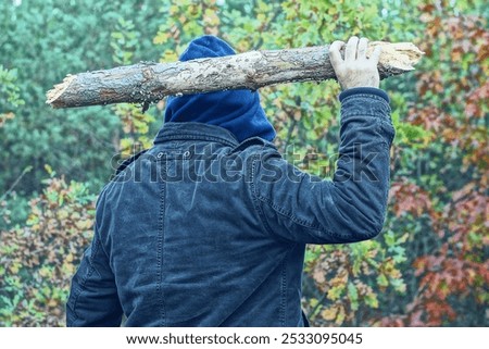 one aggressive man in a brown jacket and a blue hood on his head holds a heavy wooden wide gray stick in his hand and swings it before attacking during the day in the forest