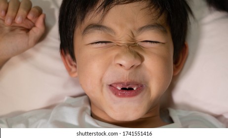 One Asian Boy Who Has The Missing Front Teeth Is Smiling And Making The Funny Face On The White Pillow When Lying Down On The Bed At Night Time.Happy Kid At Bed Time Concept.