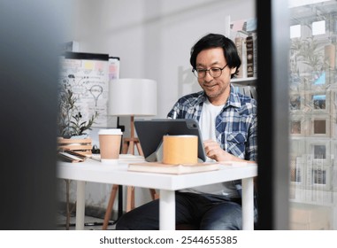 One Asian Japanese male startup worker concentrates on working with digital tablet at desk near daylight window in casual small business office, online freelance jobs, and E-commerce SME entrepreneur. - Powered by Shutterstock