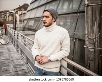 One attractive man in urban setting in modern city, leaning on metal handrail, wearing beanie cap and wool sweater, looking away