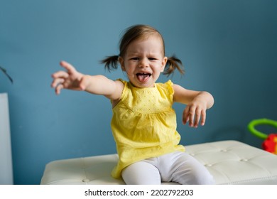 One Baby Girl Caucasian Toddler Sitting On The Couch In Front Of Blue Wall And Crying With Outstretched Arms Portrait