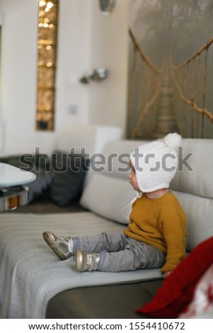 One baby in a knitted hat and sweater on the sofa, bright interior and soft focus. Concept cold at home, lack of heating