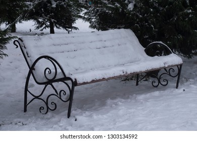 One bench in full screen. bench covered in snow. In the background is a Christmas tree. Christmas tree in the snow. It's cold outside.
