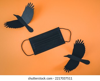 One Black Medical Mask And Two Paper Crows Lie Diagonally Against An Orange Background With Space For Text, Flat Lay Close-up. Scary Concept And Coronavirus.