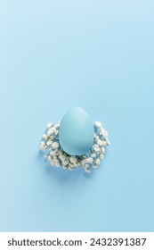 One blue Easter Egg on Bed of White Flowers on sky Blue Background. Easter celebration minimal holiday concept, top view close up pastel colored chicken egg, copy space, vertical monochrome photo