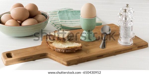 One boiled egg\
in a green eggcup with buttered toast, spoon and salt and pepper\
grinder on a wooden\
background.