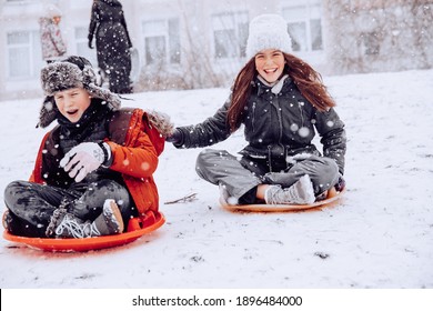 One Boy And One Girl With Long Hair On The Street In The Afternoon In Winter A Lot Of Snow Rolling Downhill On A Round Red Board Sitting Laughing In Warm Clothes Snow Falls On The Face