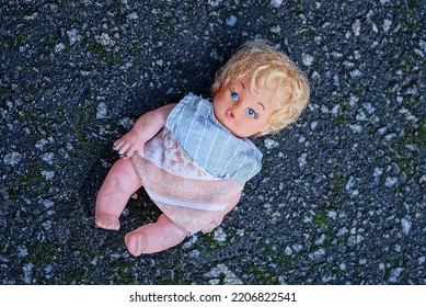 One Brown Plastic Toy Doll In A Colored Dress Lies On Gray Asphalt In The Street