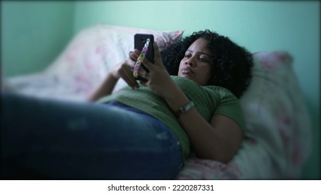 One Candid African American Woman Looking At Phone In Bed. A Black Hispanic Black Girl Texting On Smartphone Device