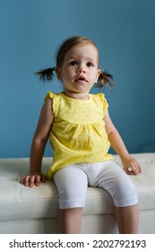 One Caucasian Baby Girl Toddler Sitting On The Couch In Front Of Blue Wall At Home Indoor Portrait Copy Space Front View