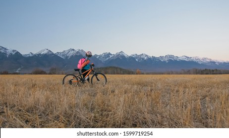 One Caucasian Children Walk With Bike In Wheat Field. Little Girl Walking Black Orange Cycle On Background Of Beautiful Snowy Mountains. Biker Stand With Backpack And Helmet. Mountain Bike Hardtail.