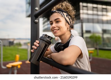 One caucasian woman taking a brake during outdoor training in the park outdoor gym resting on the bars with supplement shaker in hand drinking water or supplementation happy smile copy space - Powered by Shutterstock