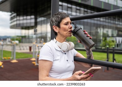 One caucasian woman taking a brake during outdoor training in the park outdoor gym resting on the bars with supplement shaker drinking water or supplementation use smartphone confident copy space