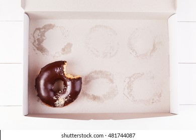 One Chocolate Bitten Donut With Sprinkles In A Empty Box On A White Wooden Background. Top View With Copyspace