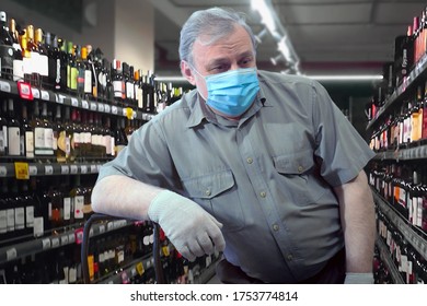 One Of The Consequences Of The Pandemic Is An Increased Craving For Alcohol. A Man In A Medical Mask And Gloves, Leaning On The Handle Of A Cart, Looks At The Rows Of Bottles Of Alcohol