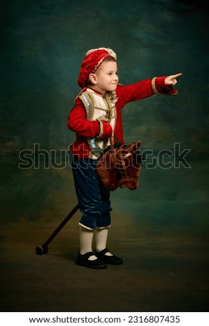 One cute little boy dressed up as medieval little prince and pageboy ride toy horse over dark vintage style background. Vintage fashion, emotions, theater art concept. Eras comparison