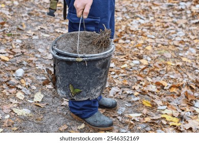 one dirty broken round old discarded black industrial open empty iron handle plastic bucket in hand of worker in blue pants daytime outdoors