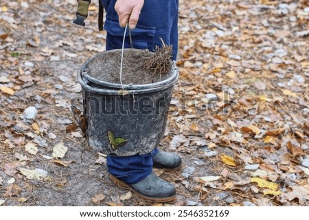 one dirty broken round old discarded black industrial open empty iron handle plastic bucket in hand of worker in blue pants daytime outdoors