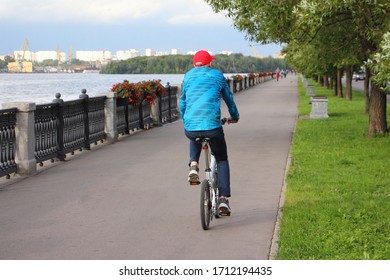 One Elderly Cyclist Man In Casual Clothing Rides On A Utility Folding Bicycle On Asphalted Quayside Alley Way On A Summer Day On Green Grass, Trees And Moscow River Background At Summer Day Rear View