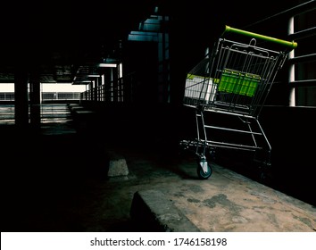 One Empty Shopping Cart Or Trolley Were Parked Lonely In Dark Parking Lot. The Cart Is In A Bright Spot Where The Light Is Come In. Perspective And Low Angle View, Space For Design Or Text.