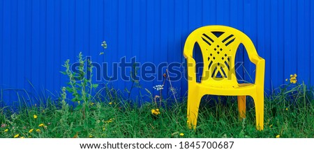 One empty yellow plastic chair standing on green grass on the background of a blue fence made of metal profile outside on summer day. Concept of outdoor vacation, relaxation, recreation, country life.