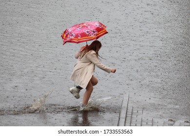 A one European girl under an umbrella in a short coat runs in white sneakers through a large puddle in heavy rain on spring day on city street - Shutterstock ID 1733553563