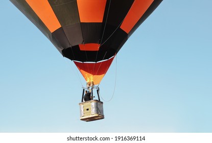 One flying striped black and orange hot air balloon with people in a basket against a clear blue sky on background.