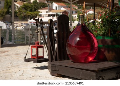 A One Gallon Glass Bottle Full Of Red Liquid On An Old Iron Scale Outside Of A Craft Shop In The KIONI Port, ITHACA, Ionian Islands, Greece In The Summer Sunny Day. Horizontal.