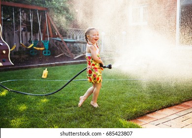 One Girl Splashing With Gardening House On Backyard On Summer Day. Child Playing With Water Outside At Sunset. Candid Moment, Lifestyle Home Kid Activity. 