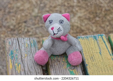 One Gray Red Plush Cat Toy Sit On A Colorful Wooden Bench Outdoors