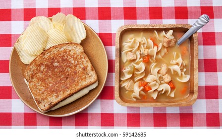 One Grilled Cheese Sandwich With Plain Chips And A Bowl Of Chicken Noodle Soup. Traditional Home Style Food On Rustic Dishes