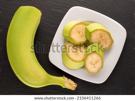 One half and four slices of an organic green banana in a white saucer on a slate stone, macro, top view.