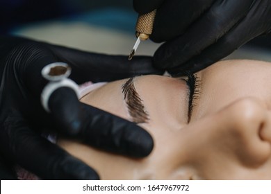 One hand in a black glove holds a manipulator with a microblading needle over the girl’s eyebrow, on the finger of the other hand is a pigment ring with a single cup closeup.