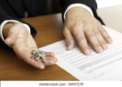 one hand of businessman gives  house keys to the hand of another businessman. Signed contract and pen visible in background.
