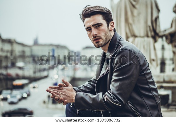 One handsome young man in urban setting in modern\
city, standing, wearing black leather jacket and jeans, looking at\
camera