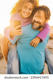 One happy couple enjoy outdoor leisure activity together with man carrying woman in piggyback and both taking a selfie with smartphone. Summer sunset in background at the beach. Travel people life - Shutterstock ID 2396417695