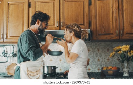 One Happy Real Couple At Home Enjoying Life Together And Relationship. Man Giving Food To Woman To Taste. Male People Cooking For His Wife. Male And Female Enjoying Leisure Activity Indoor In Kitchen